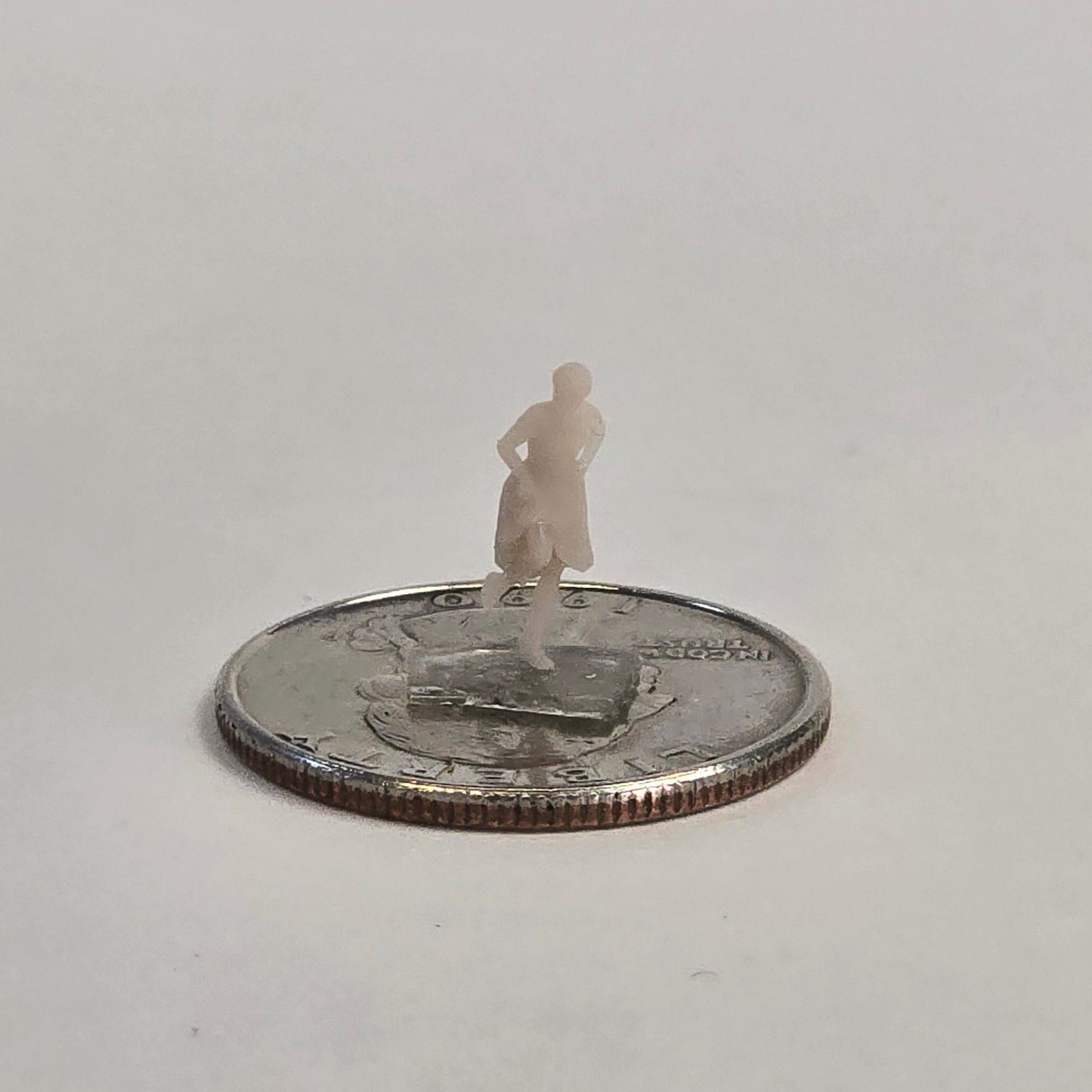 Unpainted N scale woman figure standing with hands on hips, wearing a dress and positioned on a U.S. quarter for scale reference.