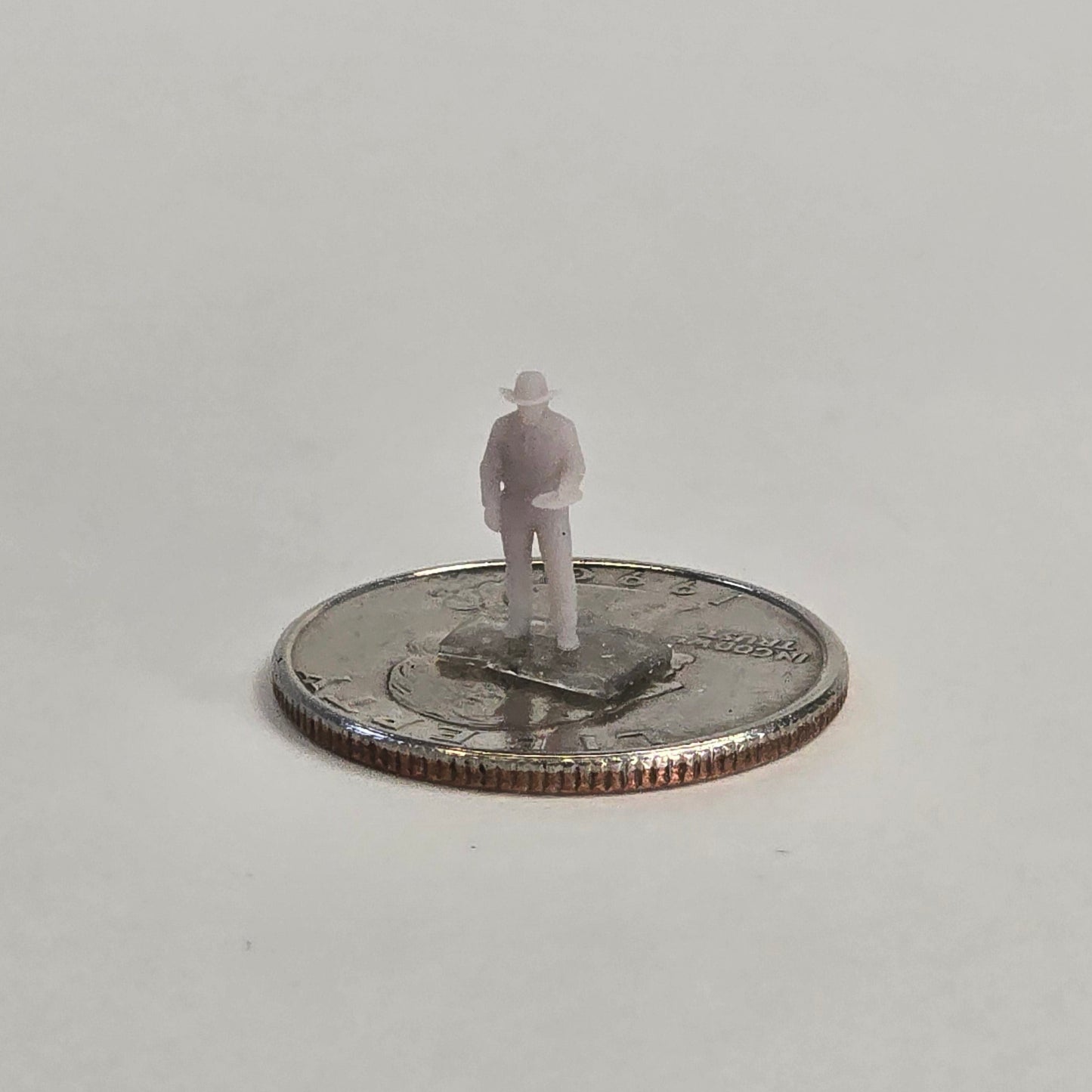 Unpainted N scale cowboy figure holding a dinner plate in his right hand, wearing a hat and standing on a U.S. quarter for scale reference.