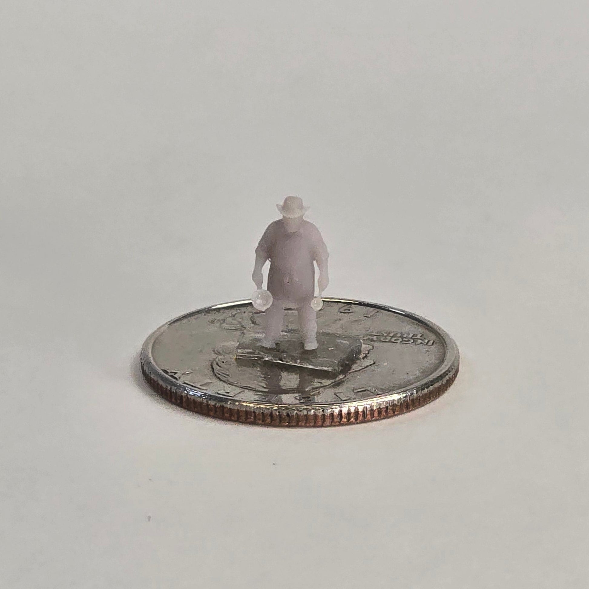 Unpainted N scale cowboy cook figure, nicknamed “Cookie,” holding a pan in each hand, wearing a hat and standing on a U.S. quarter for scale.