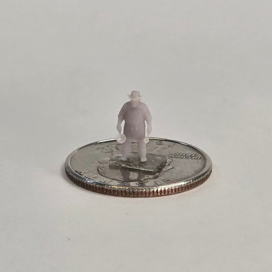 Unpainted N scale cowboy cook figure, nicknamed “Cookie,” holding a pan in each hand, wearing a hat and standing on a U.S. quarter for scale.