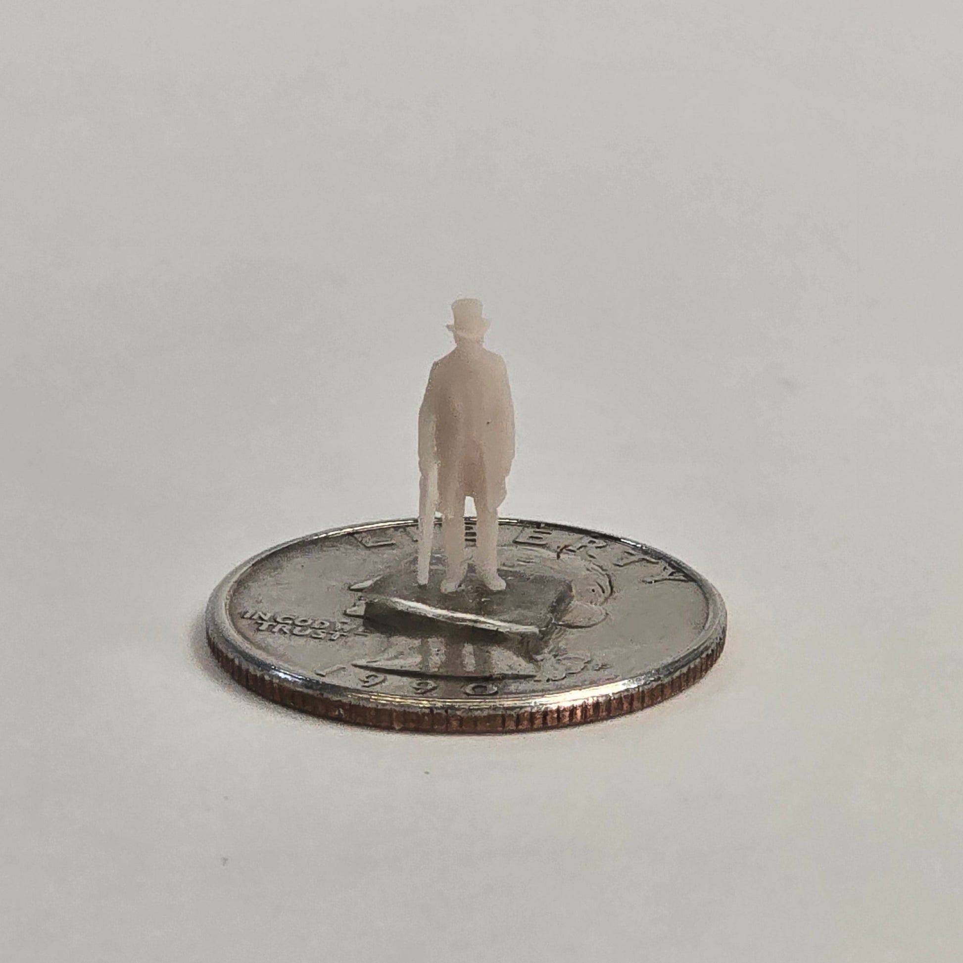 Unpainted N scale undertaker figure wearing a long coat and top hat, holding a cane, shown standing on a U.S. quarter for scale reference.