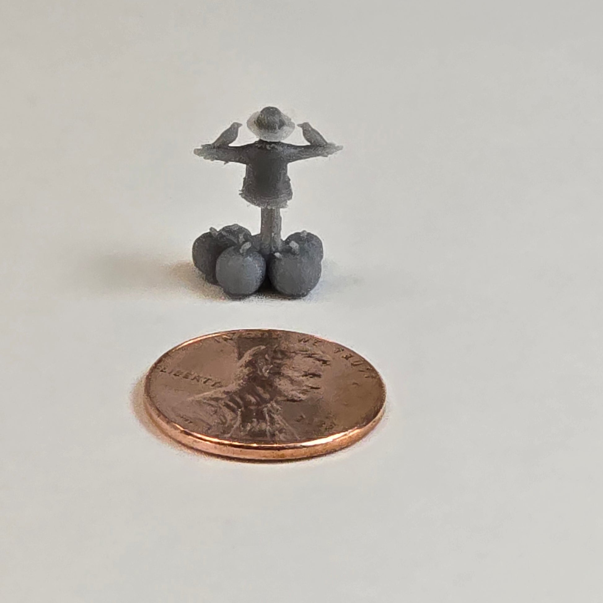 “Miniature Monday scarecrow with pumpkins — angled rear view next to a penny for scale”