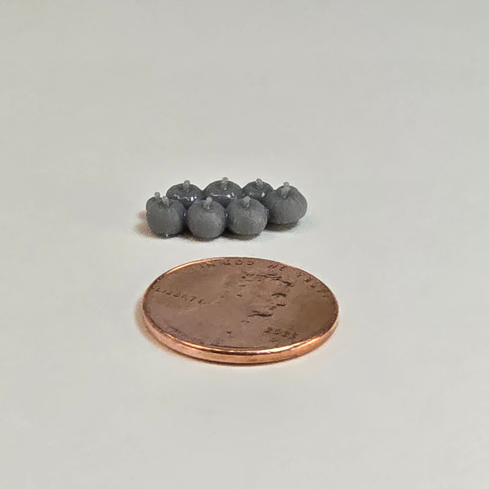 “Eight sculptural grey miniature pumpkins in two rows with a shiny penny for scale, displayed on a neutral background.”