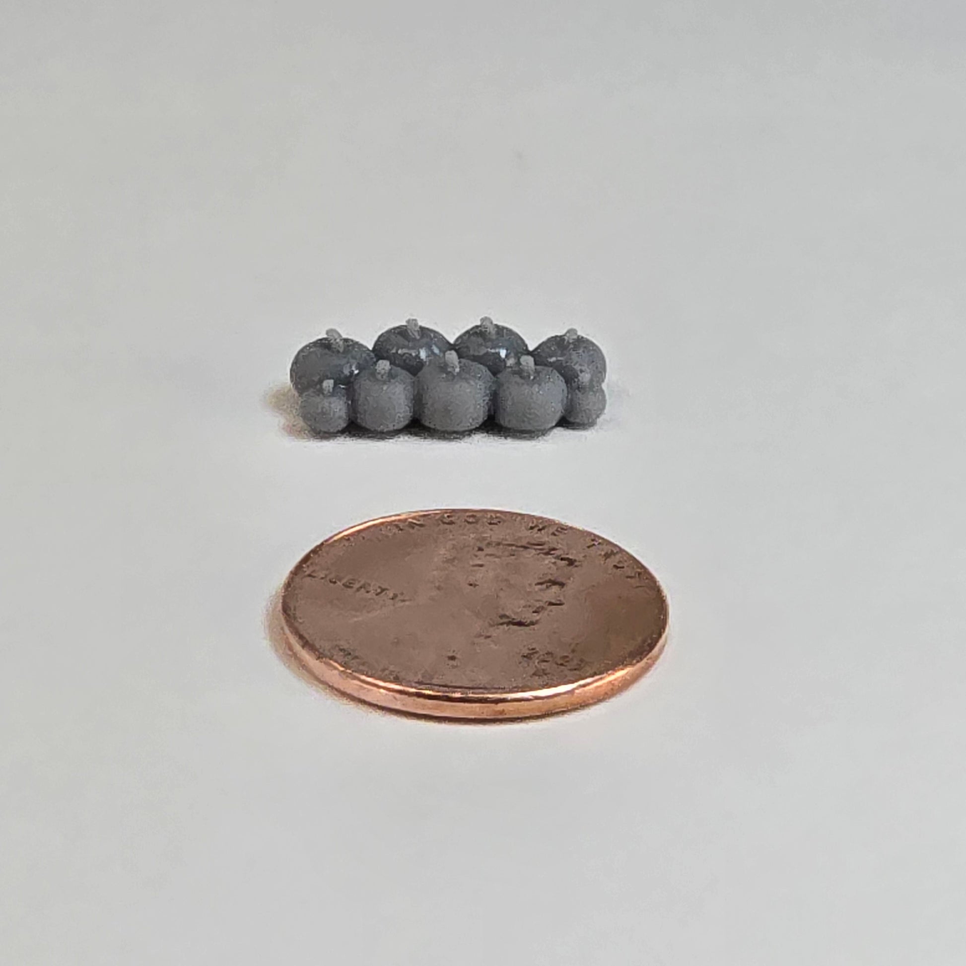 “Eight sculptural grey miniature pumpkins in two rows with a shiny penny for scale, displayed on a neutral background.”